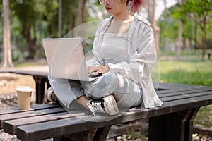A cropped image of an Asian woman sits on a bench in a public park and uses her laptop computer