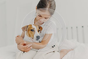 Cropped image of caring little girl in white t shirt, cuddles small pedigree dog, expresses big love to animal, poses on bed in