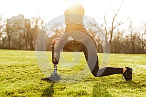 Cropped image of disabled athletic girl in black sportswear, doing lunges and stretching prosthetic leg on grass photo