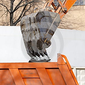 Cropped image of excavator loads the soil into the body of red dump truck, closeup