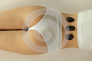 Cropped image of female body in underwear with warm stones on it isolated over grey studio background. Stone massage