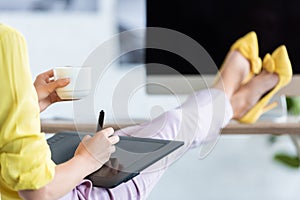 cropped image of female freelancer drinking coffee and using graphic tablet at table with computer
