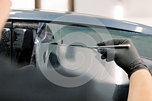 Cropped image of hands of worker in garage tinting a car window with tinted foil or film, holding special blade or knife