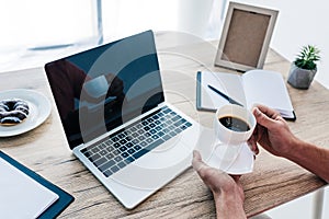 cropped image of man holding cup of coffee at table with laptop, clipboard, textbook, photo frame and place