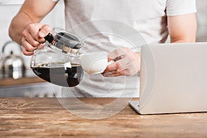 cropped image of man pouring coffee from coffee pot into cup near laptop