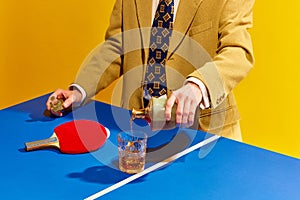 Cropped image of man in stylish suit, with tie pouring whiskey into glass on table for tennis against bright yellow