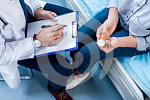 cropped image of mature male doctor writing in clipboard near female patient  with pills
