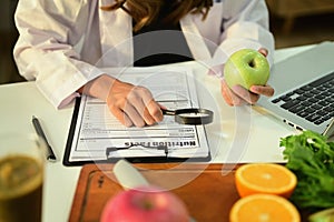 Cropped image of nutritionist holding an apple and prescribing recipe at desk with fresh fruit. Healthcare and diet
