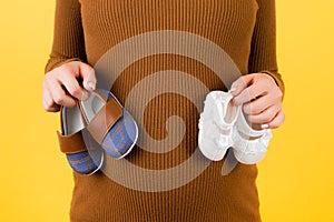 Cropped image of pregnant woman in brown dress holding two pairs of shoes for a boy and a girl at yellow background. Expecting