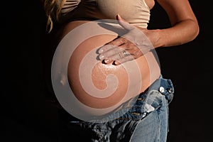 Cropped image of pregnant woman in denim jumpsuit and bra applying lotion on her belly