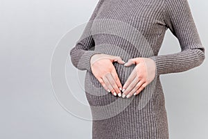Cropped image of pregnant woman holding her hands in a heart shape on her baby bump at gray background. Motherhood concept. Copy