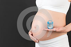 Cropped image of pregnant woman in white underwear holding alarm clock against her abdomen at black background. Child expecting.