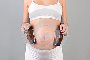 Cropped image of pregnant woman in white underwear holding booties for a baby boy against her belly at gray background. Happy