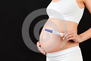 Cropped image of pregnant woman in white underwear showing positive pregnancy test against her abdomen at black background.