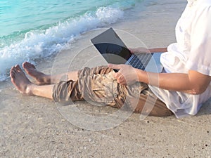 Cropped image of relaxed young Asian man with laptop sitting on sand of tropical beach in vacations day.