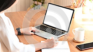 Cropped image of secretary woman`s hands while typing on white blank screen computer laptop and writing on notebook.