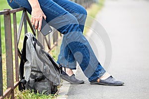 Cropped image of traveler person sitting on steel road fence, backpack is on ground, legs and hand with cellular