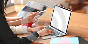 Cropped image of two beautiful women working together with computer laptop and tablet while sitting at the wooden working desk.