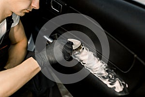 Cropped image of washing car interior by a soft brush with foam. Young male worker in black protective gloves cleaning