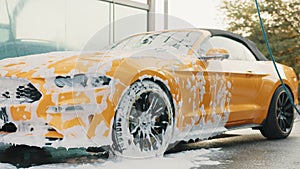 Cropped image of wheel of luxury car in outdoors self-service car wash, covered with cleaning soap foam. Washed at car