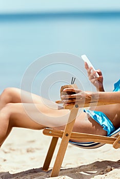 cropped image of woman holding cocktail in coconut shell and using smartphone while laying on deck chair