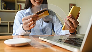 Cropped image of a woman is holding a credit card and smartphone in hands sitting in the living room.