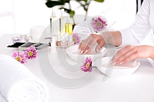 cropped image of woman receiving bath for nails at table with flowers, towels, candles, aroma oil bottles, nail polishes, cream