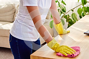 Cropped image of woman in white shirt and yellow protective rubber gloves cleaning at home and wiping dust on wooden tablel.