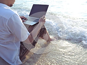 Cropped image of young relaxed man with laptop sitting on the sandy beach with soft waves. Internet of things concept.