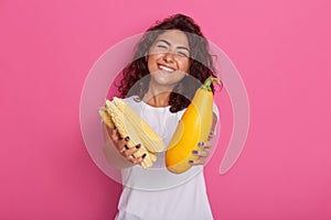 Cropped image of young woman dressed in white casual t shirt showing zucchini and corncob to camera, has happy facial expresion, photo