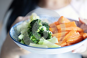 Cropped image of young woman dressed in white t-shirt showing broccoli and carrot in a plate to camera