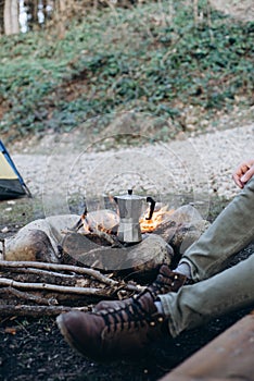 Cropped outdoor image of young man explorer sitting near to bonfire preparing hot beverage in mountains. Concept adventure