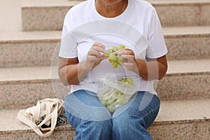 cropped photo of adult woman on stairs and eating grapes on summer warm day. female in casual clothes eating fresh green