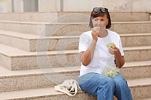 cropped photo of adult woman on stairs and eating grapes on summer warm day. female in casual clothes eating fresh green
