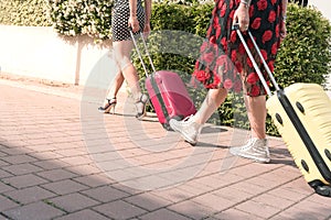 Cropped photo from below of two sexy tourist women walking with suitcases on the flowery street of the city