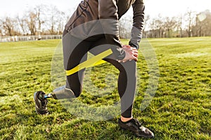 Cropped photo of disabled girl in tracksuit, exercising and doing lunges with prosthetic leg on grass using resistance band