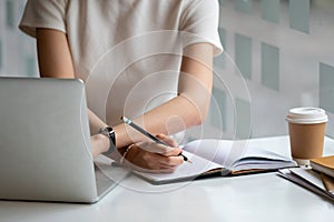 Cropped photo hand of woman writing making list taking notes in notepad working or learning online with laptop at home