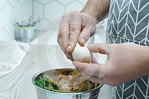 Cropped photo of man wearing grey apron peeling hardboiled egg over metal bowl full of carrot onion peelings. Recycling.