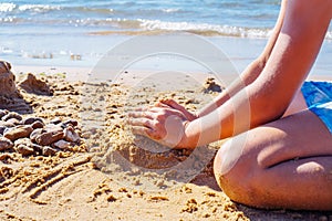 Cropped photo of preteen boy sitting on beach seashore coastline, playing with wet sand building castles near sea waves.