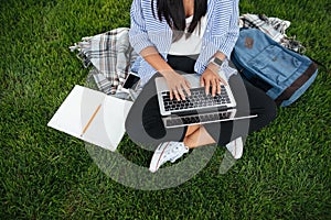 Cropped photo of student girl in casual wear, using laptop, sitting on grass, outdoor