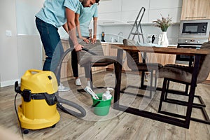 Cropped photo of two young professional cleaners in uniform using vacuum cleaner while cleaning kitchen chairs. Cleaning
