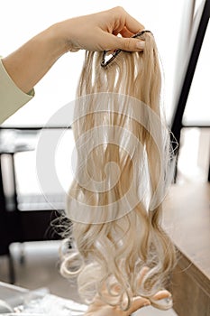 Cropped photo of woman hairdresser holding long fair blond hairpiece chignon weave bundle for extension in beauty salon.