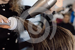 Cropped photo of woman with long dark hair sitting in salon. Hairstylist making hairstyle using dryer metal round brush.