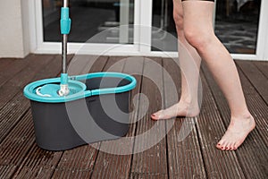 Cropped photo of young barefoot woman holding spin mop over grey plastic bucket, cleaning wooden floor of veranda.