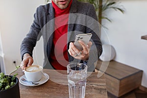 Cropped photo of young handsome unshaved man dressed in red roll-neck sweater and grey woolen jacket holding mobile phone in