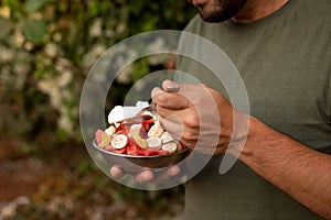 Cropped photo of young man holding tropical fruit salad with cut various exotic fruits metal bowl, fork, eating outside.