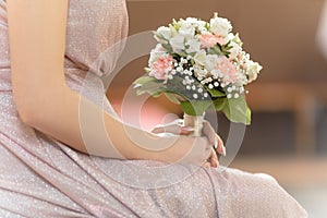 Cropped photo of young woman bride wearing long pink wedding dress, sitting holding bridal bouquet of white, pink roses.