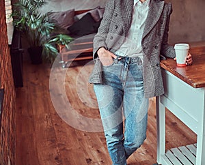 Cropped photo of a young woman dressed in a gray elegant jacket and jeans holding a cup of takeaway coffee while leaning