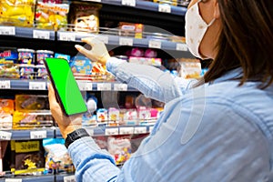 Cropped photo of a young woman with face mask using mobile phone and buying groceries in the supermarket during virus