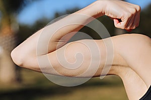 Cropped photo of young woman& x27;s arm with muscles, biceps on nature background. Girl close up of demonstrates strong